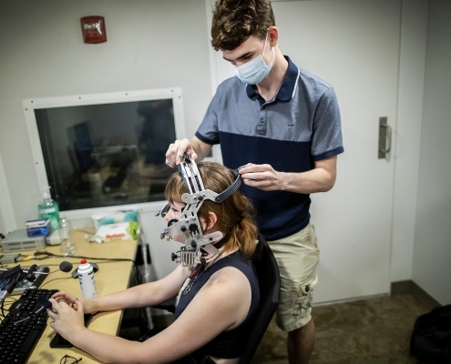 A person in a mask standing, putting a headset on a person sitting at the desk.